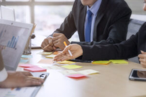 Personen im Anzug sitzen an einem Konferenztisch, auf dem bunte Haftnotizen, Laptops und Diagramme liegen. Eine Hand zeigt auf die Notizen, während diskutiert wird.