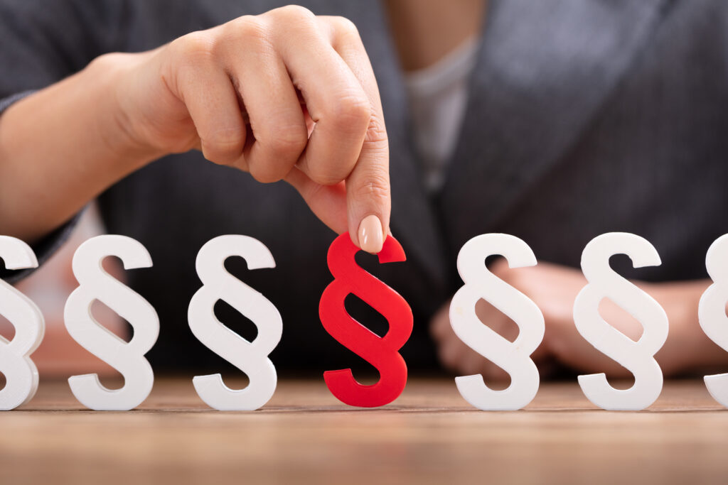 Eine Person greift nach einem roten Paragrafenzeichen, das zwischen mehreren weißen Paragrafensymbolen aufgestellt ist.