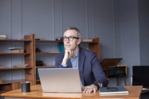 Ein Mann mittleren Alters mit Brille sitzt nachdenklich an einem Schreibtisch vor einem Laptop. Er stützt sein Kinn auf die Hand und blickt zur Seite. Auf dem Tisch liegen außerdem ein Notizbuch und eine Kaffeetasse.