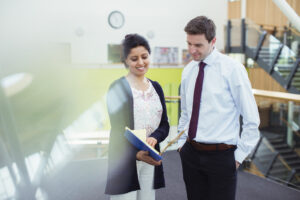 Eine Frau und ein Mann stehen in einem modernen Bürogebäude und betrachten gemeinsam ein geöffnetes Dokument in einer blauen Mappe. Beide lächeln und wirken konzentriert im Gespräch.