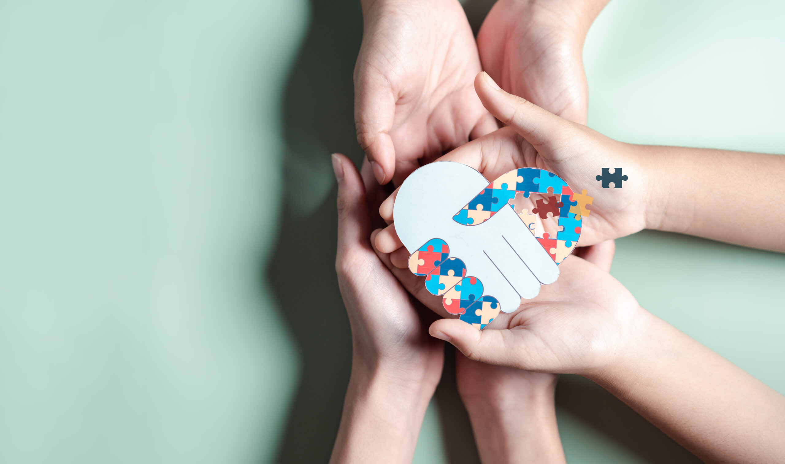 Mehrere Hände halten ein Herz aus bunten Puzzleteilen mit einem Handschlag-Motiv.