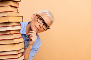 Eine Frau mit kurzen grauen Haaren und Brille schaut neugierig hinter einem hohen Stapel Bücher hervor. Sie trägt ein blau gestreiftes Hemd und hält nachdenklich ihre Hand ans Kinn.