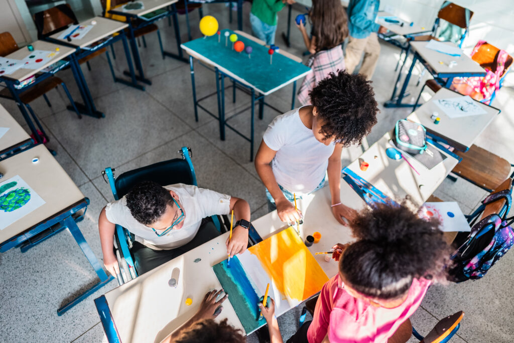 Mehrere Grundschulkinder sitzen und stehen an Tischen in einem Klassenzimmer und malen bunte Bilder. Auf den Tischen liegen Farben, Papier und Malutensilien. Die Szene zeigt kreatives Arbeiten in Gruppen.