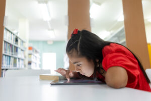 Kind in rotem Kleid arbeitet in einer Bibliothek konzentriert an einem Tablet auf dem Tisch.