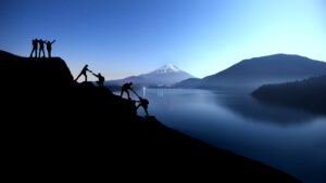 Silhouetten von Menschen, die sich gegenseitig beim Aufstieg auf einen Berg helfen, im Hintergrund ein See und ein schneebedeckter Gipfel.