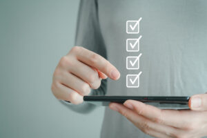 Person hält ein Smartphone und tippt darauf, daneben sind digitale Häkchen in Kästchen eingeblendet.