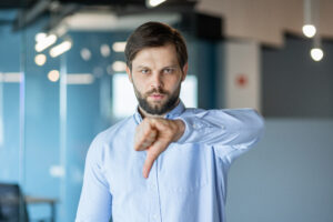 Ein Mann mit Bart und hellem Hemd steht in einem modernen Büro und zeigt mit ernstem Gesichtsausdruck den Daumen nach unten.