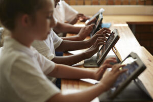 Mehrere Schülerinnen und Schüler sitzen im Klassenzimmer und arbeiten konzentriert mit Tablets. Die Kinder tragen weiße Oberteile, die Tablets stehen in Schutzhüllen mit Ständern auf den Holztischen.