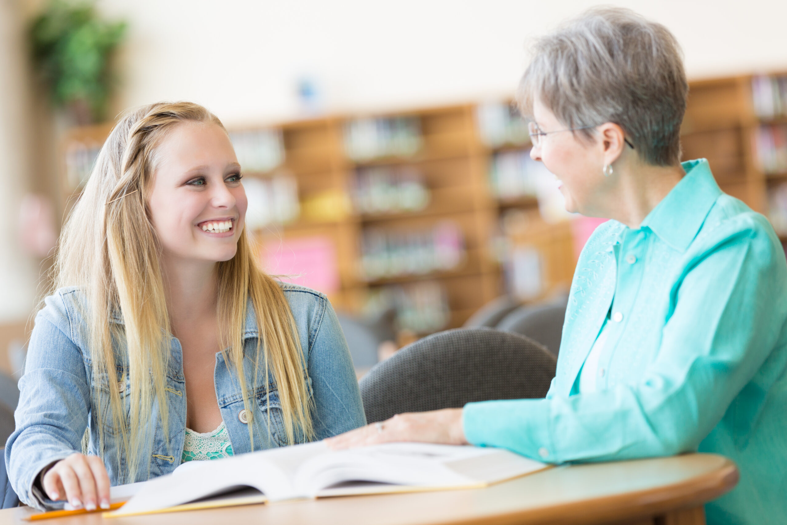 Eine ältere Frau und eine junge Schülerin sitzen zusammen an einem Tisch in einer Bibliothek. Vor ihnen liegt ein aufgeschlagenes Buch, beide unterhalten sich lächelnd.