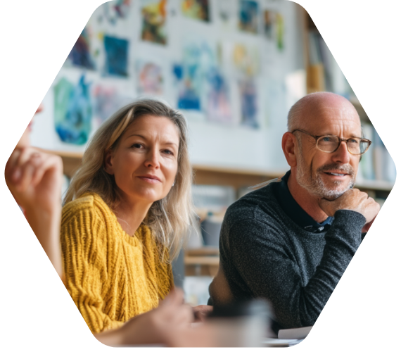 Zwei Erwachsene, eine Frau im gelben Pullover und ein Mann mit Brille, sitzen aufmerksam in einer Gesprächssituation, im Hintergrund hängen bunte Bilder an der Wand.