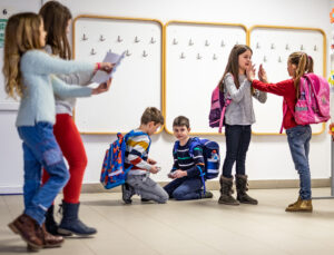Mehrere Grundschulkinder mit Schulranzen befinden sich im Flur einer Schule. Zwei Mädchen interagieren direkt miteinander, zwei Jungen knien am Boden und beschäftigen sich mit etwas, während zwei weitere Mädchen mit Zetteln in der Hand vorbeigehen.