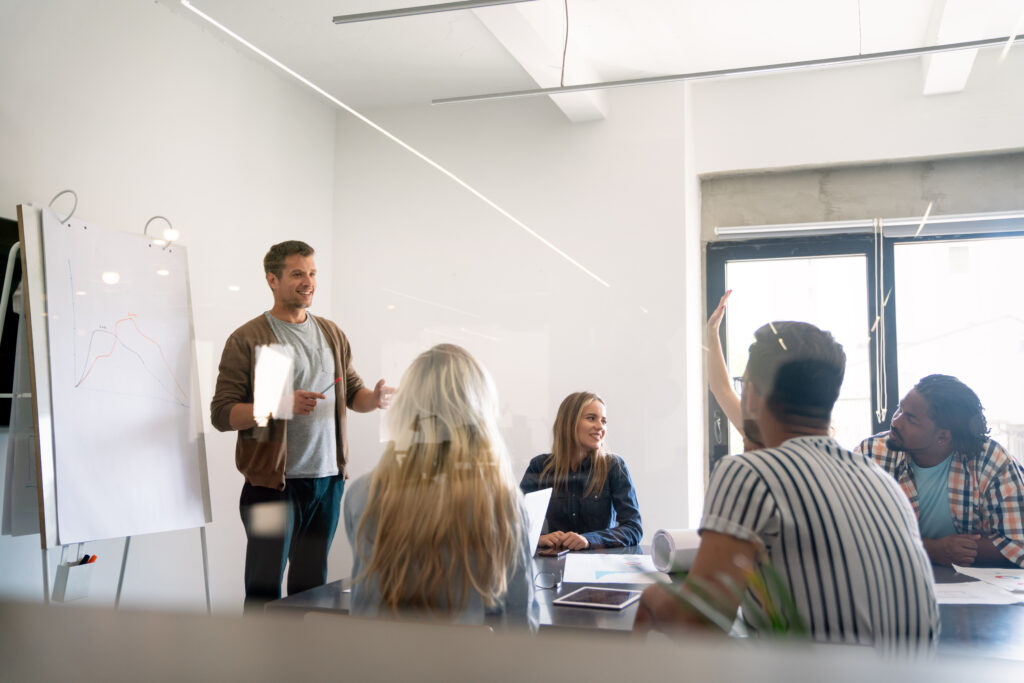 Ein Mann steht vor einem Flipchart mit Diagramm und spricht zu einer Gruppe von fünf Personen, die an einem Tisch sitzen.