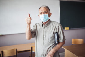 Ein älterer Lehrer steht im Klassenzimmer, trägt eine medizinische Schutzmaske und hebt den Daumen nach oben. Unter dem Arm hält er ein Notizbuch. Im Hintergrund sind Tische, Stühle und eine Tafel zu sehen.