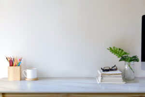 Aufgeräumter Schreibtisch mit Stiftehalter, Tasse, Büchern, Brille und einer Pflanze im Glasgefäß.