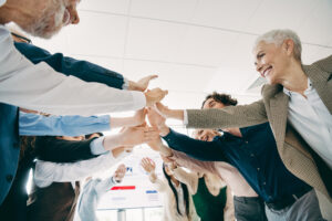 Eine Gruppe von Menschen steht im Kreis und streckt ihre Hände in die Mitte. Alle legen die Hände übereinander, lächeln und wirken motiviert und verbunden.