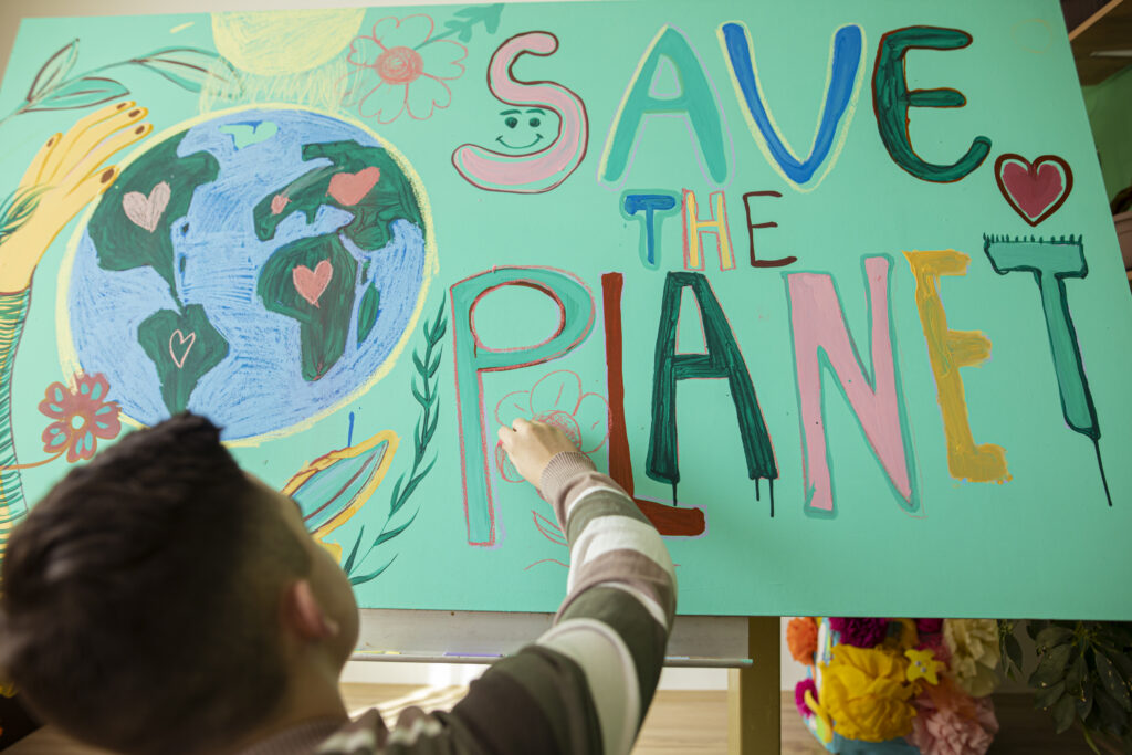 Ein Kind malt an einem farbenfrohen Plakat mit der Aufschrift „SAVE THE PLANET“ und einer Zeichnung der Erde.