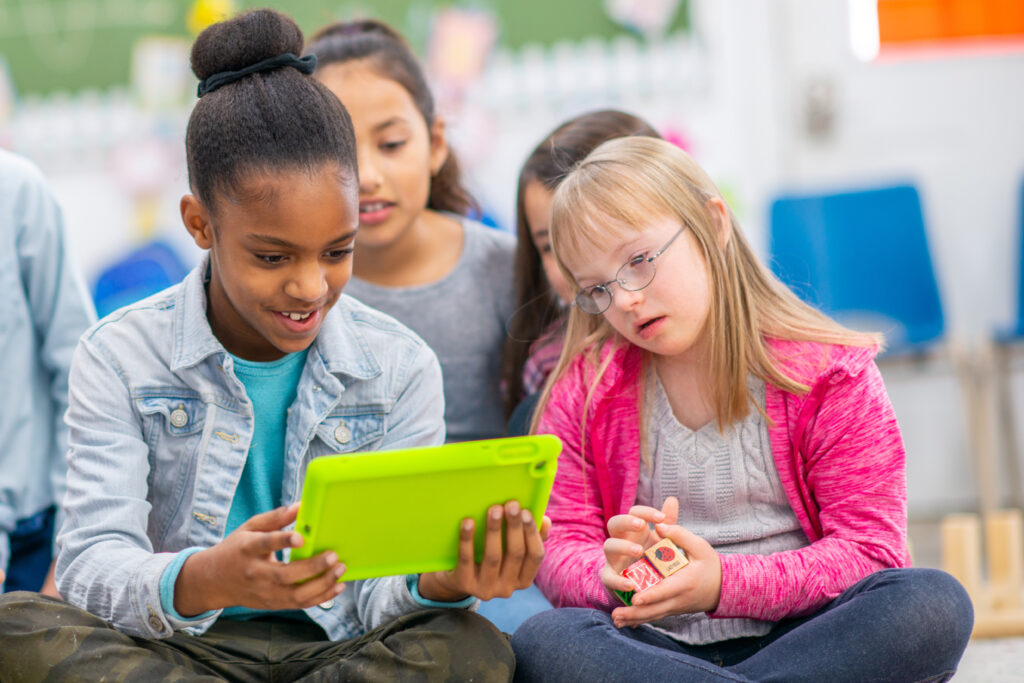 Eine kleine Gruppe von Grundschulkindern drängt sich um ein Tablet und sieht sich eine elektronische Unterrichtsstunde an. Sie sind alle leger gekleidet und konzentrieren sich auf den Bildschirm.