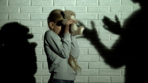 Ein verängstigtes Mädchen hält einen Teddybär schützend vor ihr Gesicht, während die Schatten von Erwachsenen drohend an der Wand erscheinen.