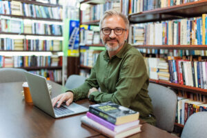 Ein lächelnder Mann mit Brille sitzt in einer Bibliothek am Tisch, arbeitet an einem Laptop und ist von Büchern umgeben.