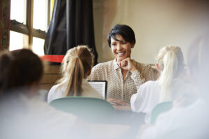 Lehrerin sitzt an einem Tisch und lächelt Schülerinnen an, die mit digitalen Geräten arbeiten.