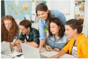 Eine Lehrerin arbeitet gemeinsam mit vier Schülerinnen und Schülern an Laptops und Büchern. Alle sitzen um einen Tisch und sind konzentriert in eine Aufgabe vertieft.