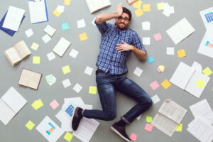 Mann liegt gestresst am Boden, umgeben von vielen Zetteln, Büchern und Dokumenten.