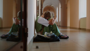 Mädchen sitzt allein im Schulflur auf dem Boden, mit angezogenen Knien und gesenktem Kopf.