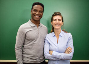 Zwei Personen stehen vor einer grünen Tafel, eine Person legt der anderen den Arm auf die Schulter. Beide lächeln in die Kamera.