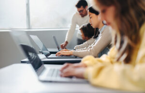 Gruppe von Schulkindern arbeitet mit digitalen Geräten wie Laptops und Tablets im Klassenzimmer, begleitet von einer Lehrkraft.