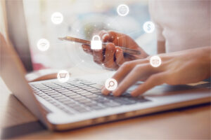 Eine Person tippt auf einem Laptop und hält gleichzeitig ein Smartphone in der Hand. Um das Smartphone sind Symbole wie Schloss, Einkaufswagen, E-Mail, Lupe, WLAN und Dollarzeichen eingeblendet.