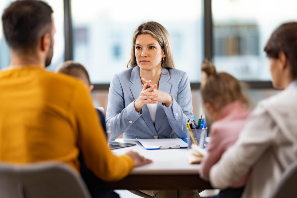 Frau in hellblauem Blazer sitzt an einem Tisch und hört konzentriert einer Familie zu, die ihr gegenübersitzt.