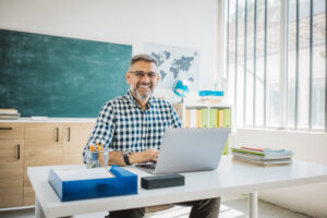 Ein Mann mit Brille und kariertem Hemd sitzt an einem Schreibtisch mit Laptop und schaut in die Kamera.