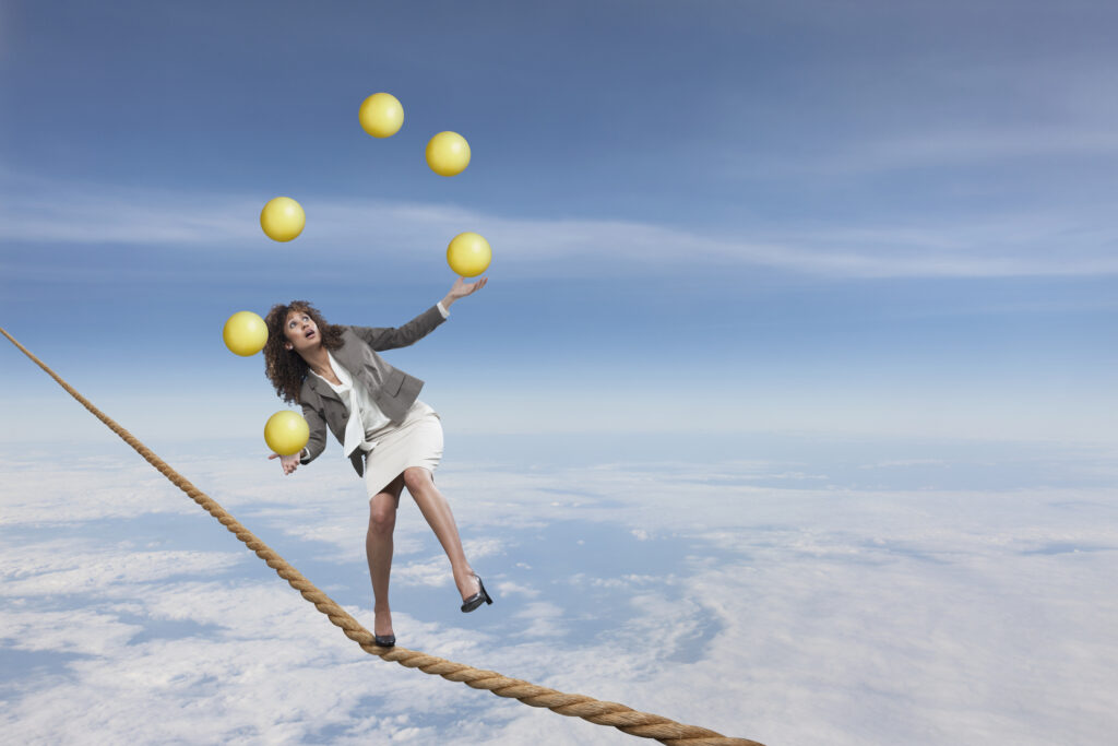 Eine Frau im Business-Outfit balanciert auf einem Seil über den Wolken und jongliert gleichzeitig mit mehreren gelben Bällen.
