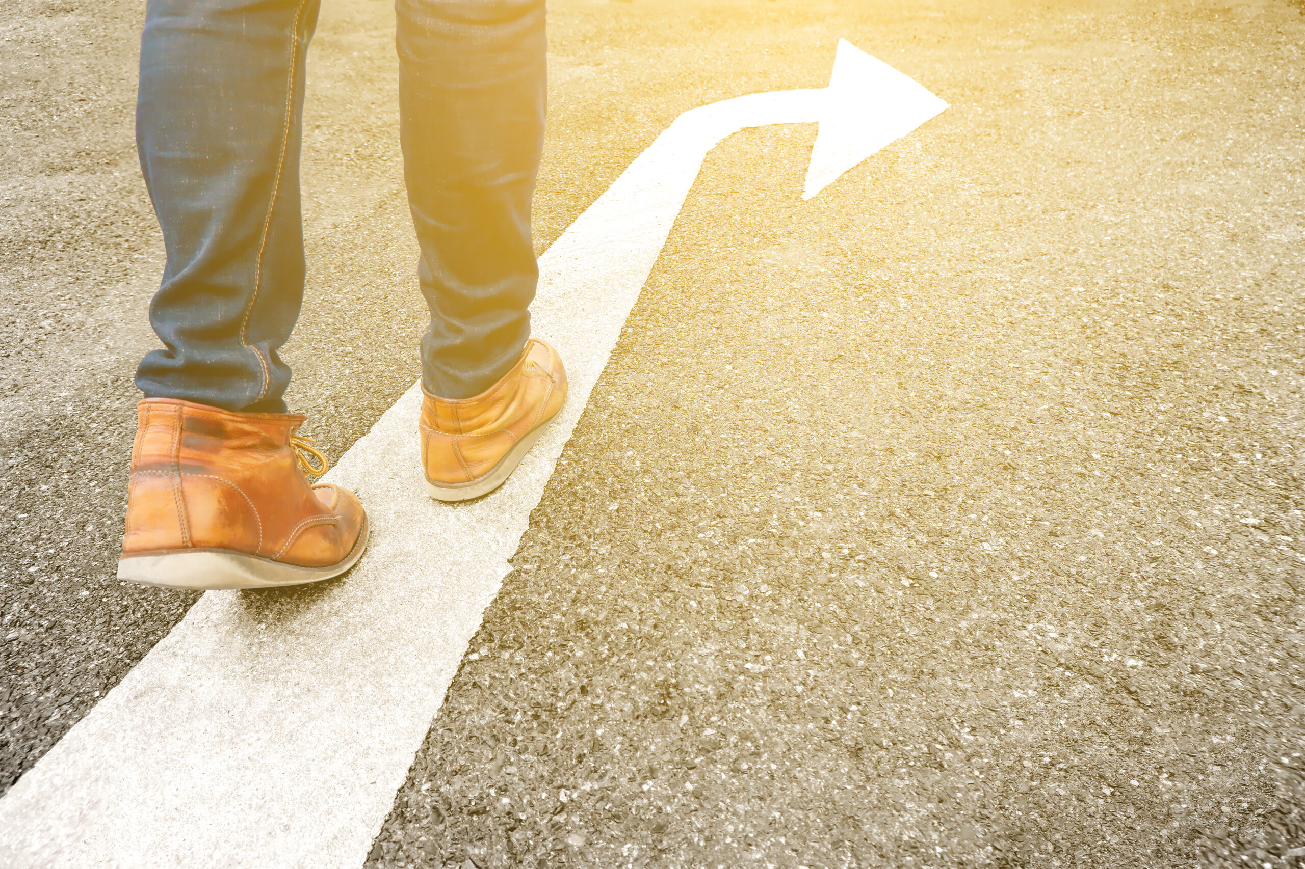 Person mit braunen Lederschuhen geht auf einer weißen Linie auf einer Asphaltstraße, im warmen Sonnenlicht.