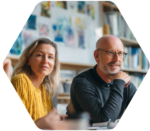 Zwei erwachsene Personen – eine Frau in gelbem Pullover und ein Mann mit Brille in dunklem Pullover – sitzen aufmerksam in einem hellen Raum mit Bücherregalen und farbigen Bildern an der Wand. Sie wirken konzentriert und interessiert, als würden sie an einer Besprechung oder Weiterbildung teilnehmen.