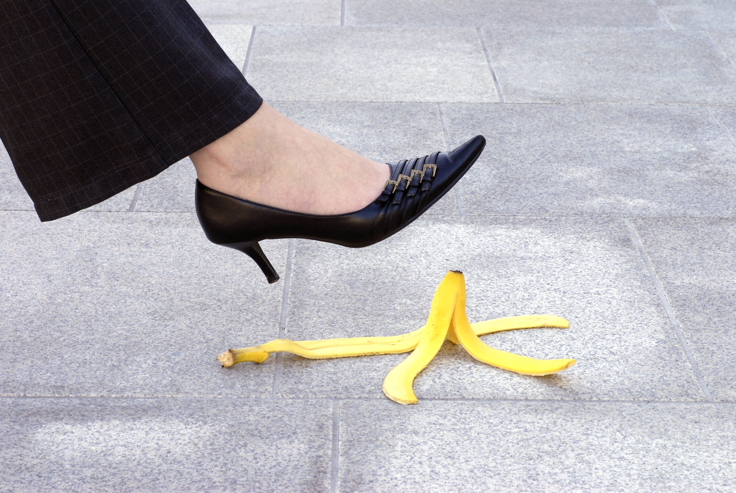 Ein Fuß in einem schwarzen Absatzschuh steht kurz davor, auf eine Bananenschale auf dem Gehweg zu treten.