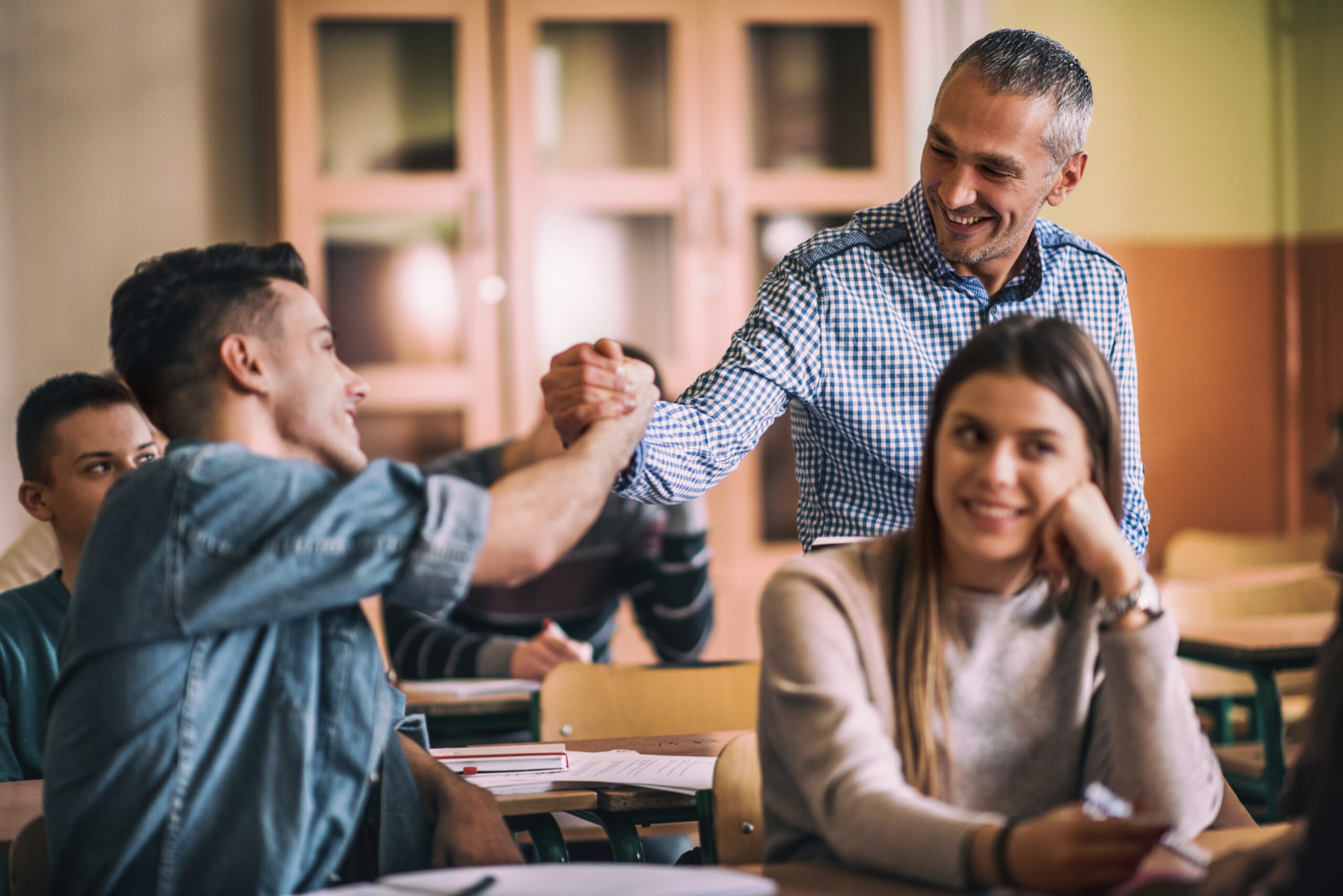 Ein Lehrer begrüßt einen Schüler in einem Klassenraum mit einem freundlichen Handschlag, während andere Schüler im Hintergrund lächeln und zusehen. Ein Lehrer begrüßt einen Schüler in einem Klassenraum mit einem freundlichen Handschlag, während andere Schüler im Hintergrund lächeln und zusehen.