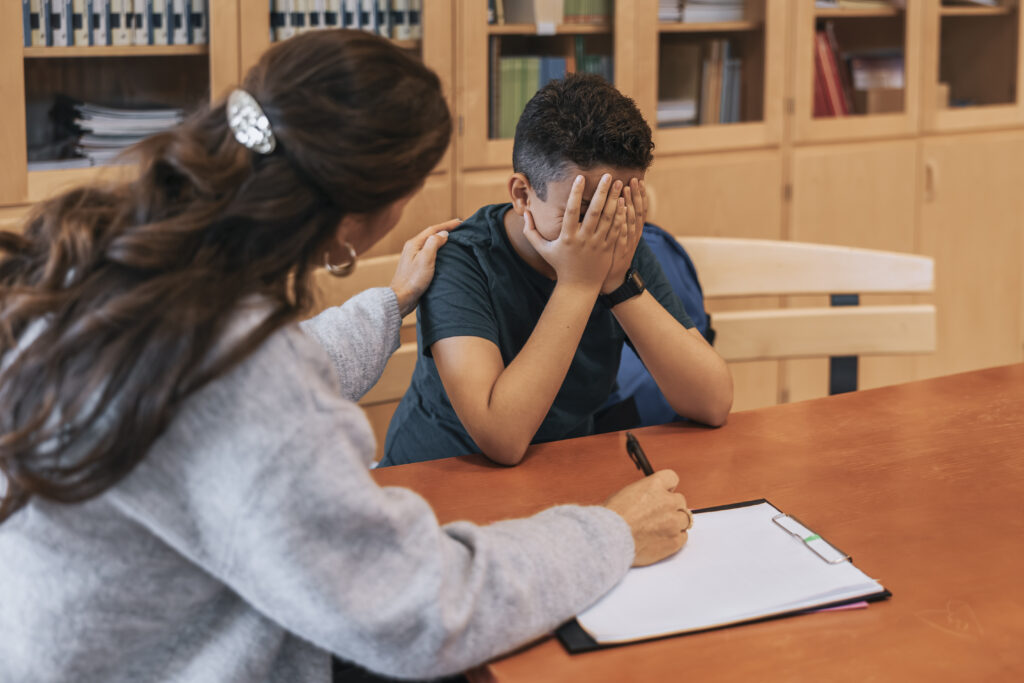 Lehrerin spricht mit einem Schüler, der sein Gesicht in den Händen verbirgt, und legt beruhigend eine Hand auf seine Schulter.