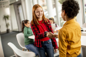 Zwei Kinder geben sich freundlich die Hand, während andere Kinder im Hintergrund im Klassenraum arbeiten.