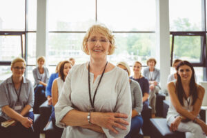 Gruppe von Lehrkräften, im Vordergrund eine lächelnde Person, die vor der sitzenden Gruppe steht.