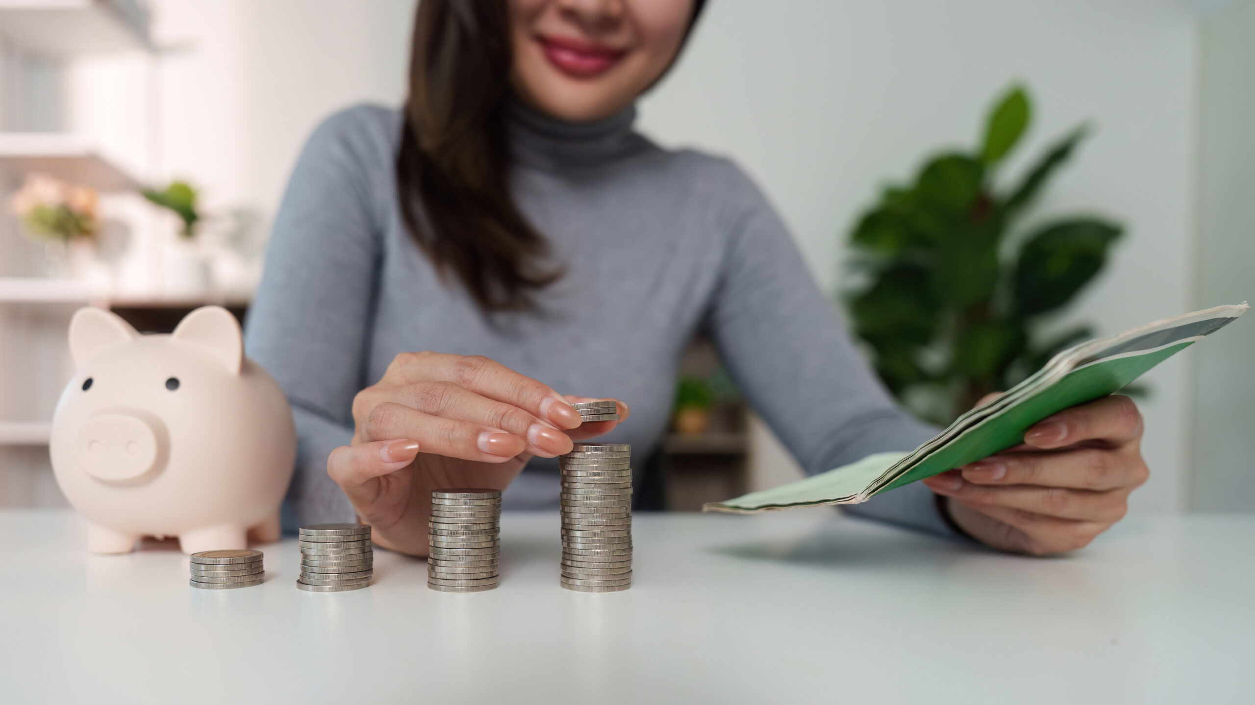 Person stapelt Münzen auf dem Tisch, neben einem Sparschwein; in der Hand hält sie ein Notizbuch zur Budgetplanung.