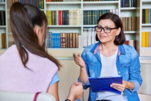 Frau im Gespräch mit einer jüngeren Frau in einem Büro; sie hält ein Klemmbrett.