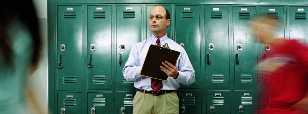 Ein Schulinspektor mit Krawatte und Klemmbrett steht in einem Schulflur vor einer Reihe grüner Schließfächer. Links und rechts laufen unscharf Schülerinnen und Schüler vorbei, wodurch Bewegung im Bild entsteht.