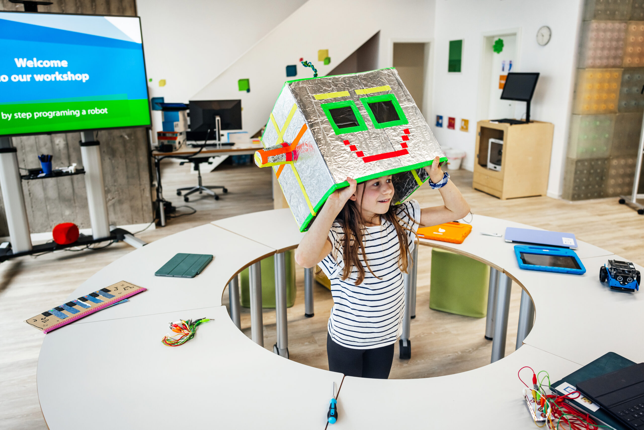Ein Mädchen steht in einem hellen Workshop-Raum und trägt einen selbstgebastelten silbernen Roboterhelm aus Pappe mit grün umrandeten Augen und rotem Mund. Um sie herum stehen Tische mit Tablets, Kabeln und einem kleinen Roboterauto; links zeigt ein Bildschirm eine Willkommensfolie für den Workshop.