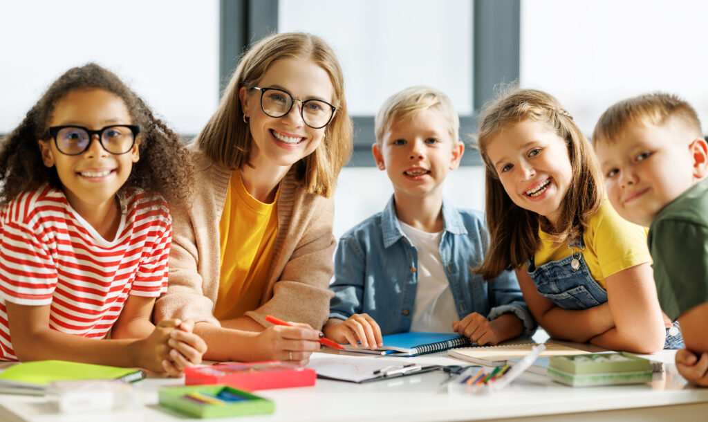 Lehrerin sitzt mit vier Grundschulkindern an einem Tisch mit Heften und Stiften in einem hellen Klassenraum und alle lächeln in die Kamera.