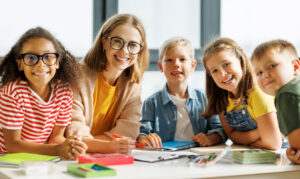 Lehrerin sitzt mit vier Grundschulkindern an einem Tisch mit Heften und Stiften in einem hellen Klassenraum und alle lächeln in die Kamera.