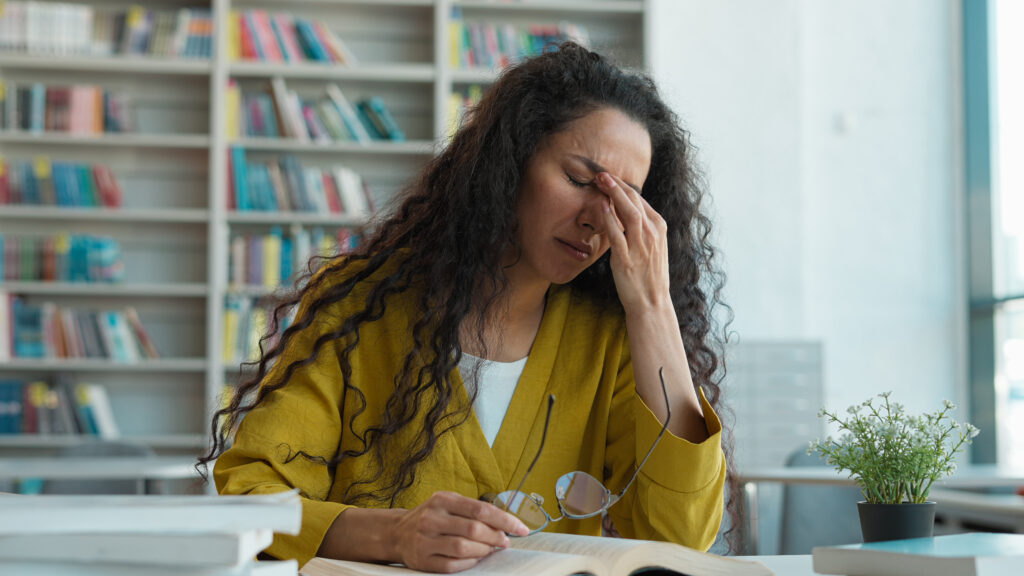 Eine Lehrerin sitzt gestresst an ihrem Schreibtisch und reibt sich erschöpft die Augen.
