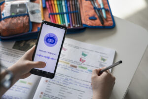 Kind hält Smartphone mit KI-Assistenz-App, während es handschriftlich in einem Schulheft arbeitet; Stiftebox im Hintergrund.