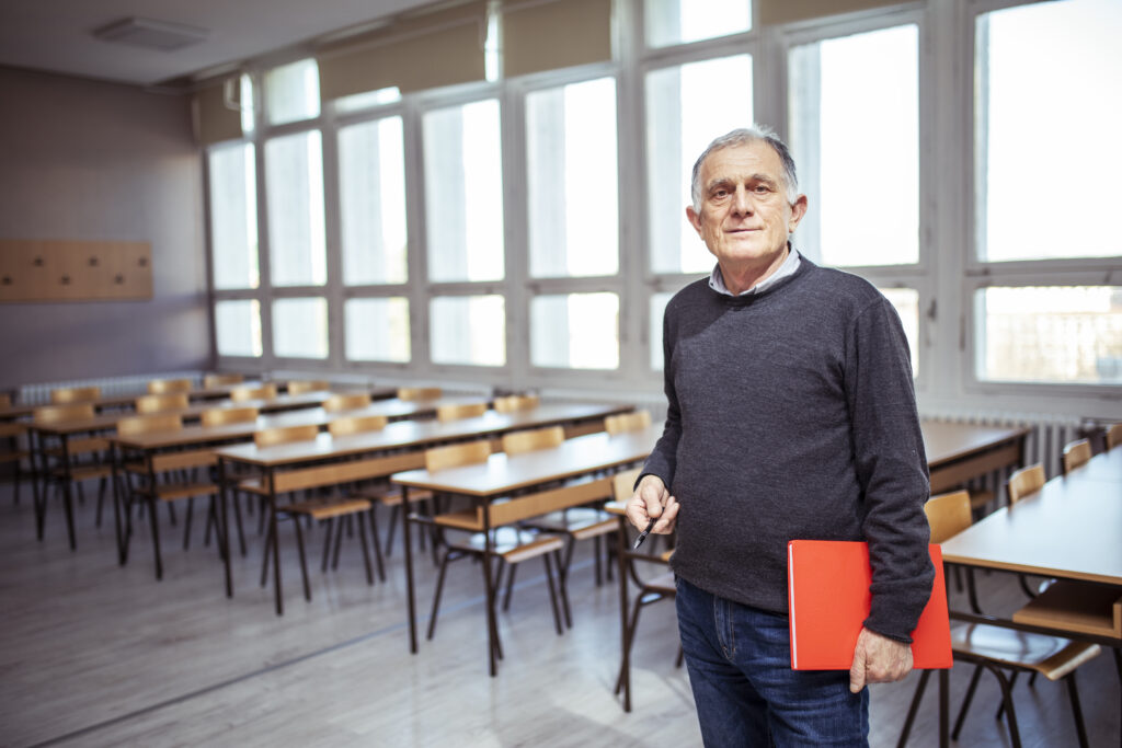 Älterer Lehrer mit roter Mappe und Stift in einem leeren Klassenzimmer